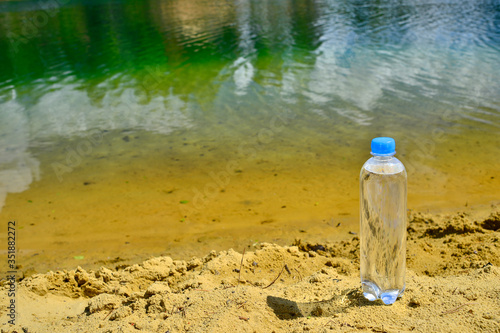 A closed plastic water bottle with a close up stands on a sandy beach against the background of clear lake water.