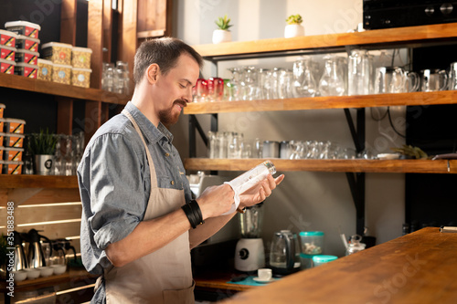 Happy young bearded waiter in apron wiping glass with napkin or towel