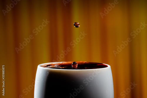 Red wine splash. Reaction of a falling drop on a red wine  surface in a completely filled white plastic cup. Studio photo with matching orange striped colored background. Beautiful wine drop.