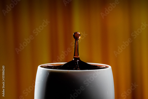 Red wine splash. Reaction of a falling drop on a red wine  surface in a completely filled white plastic cup. Studio photo with matching orange striped colored background. Beautiful wine drop.