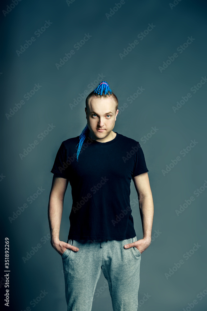 A man with a braided mohawk haircut calmly posing holding hands in pockets of sweatpants