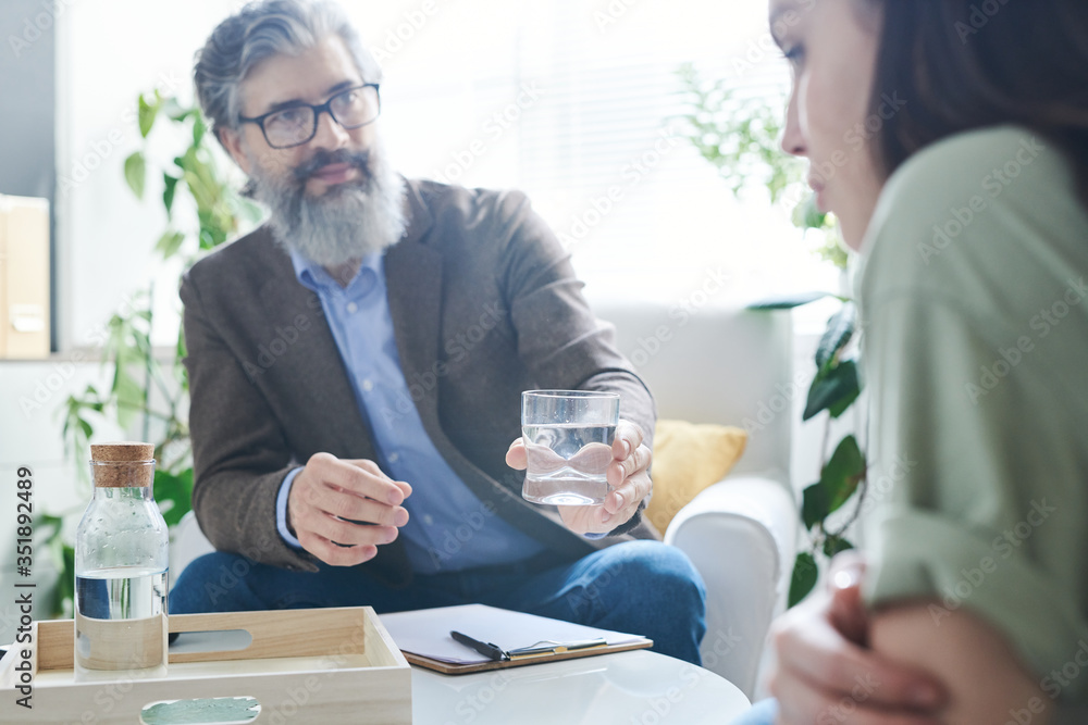 Senior counselor with beard passing glass of water to his young female patient Senior counselor with beard passing glass of water to his young female patient