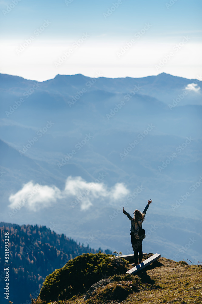 Sunny mountain view. Young woman with a long hair and with a backpack raised her hands up in the air. She is looking to the horizon and greeting the sky and the mountains on the background. 