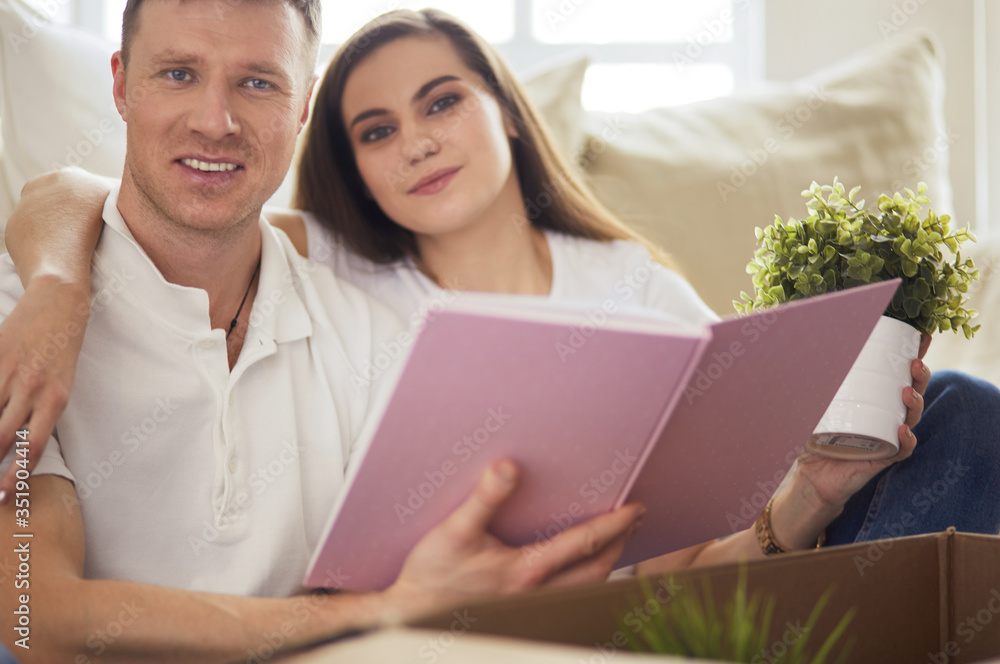 Cute couple unpacking cardboard boxes in their new home, sitting on the floor and looking at a family album Cute couple unpacking cardboard boxes in their new home, sitting on the floor and looking at a family album