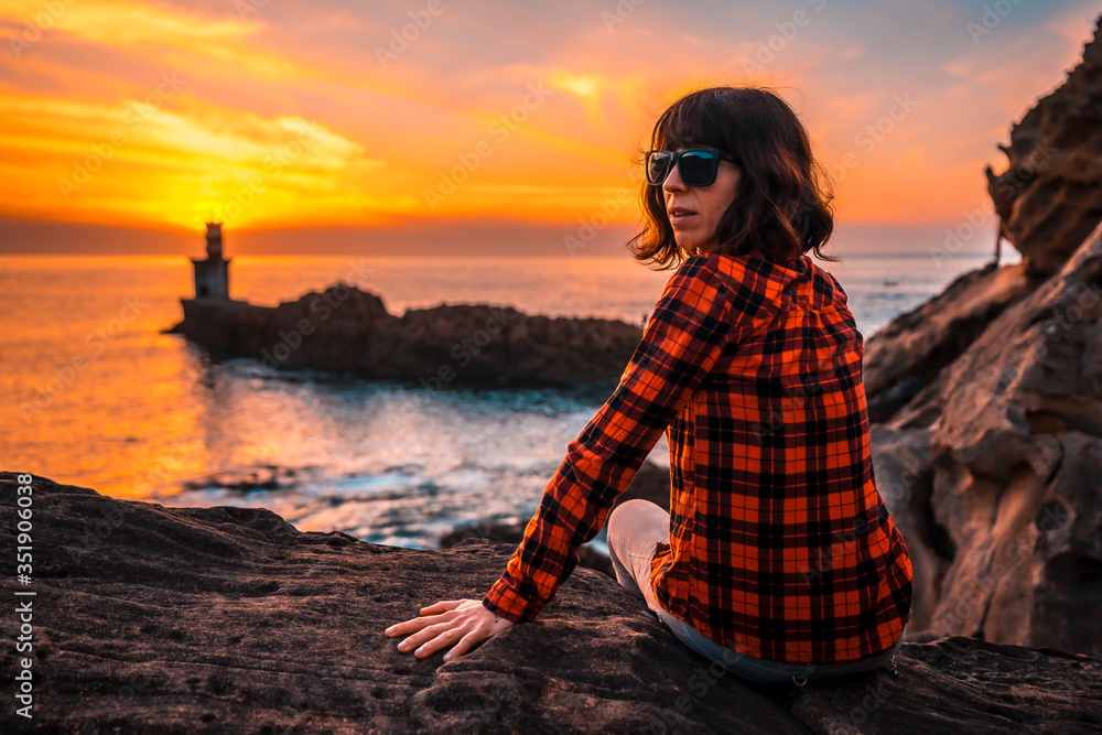 A young girl sitting with sunglasses in the orange sunset of the Pasajes San Juan lighthouse, near San Sebastian, Gipuzkoa. Basque Country