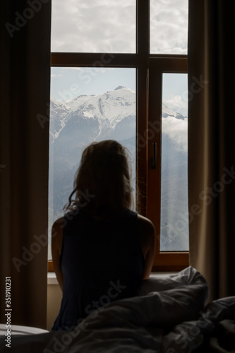 A girl sits on a bed in her room in front of the window and admires the morning mountain landscape. She delight in view of snow-capped peaks and green forests of the Caucasus Mountains.