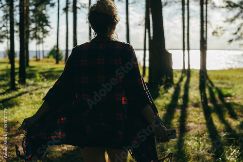A girl in a checkered poncho runs to meet a sparkling river visible among pine trees © Anton