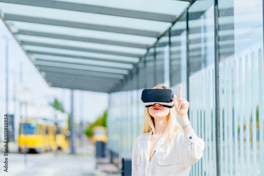Young woman with vr goggle pushing virtual button while standing against glass modern bus stop outdoor. Sun glare effect.