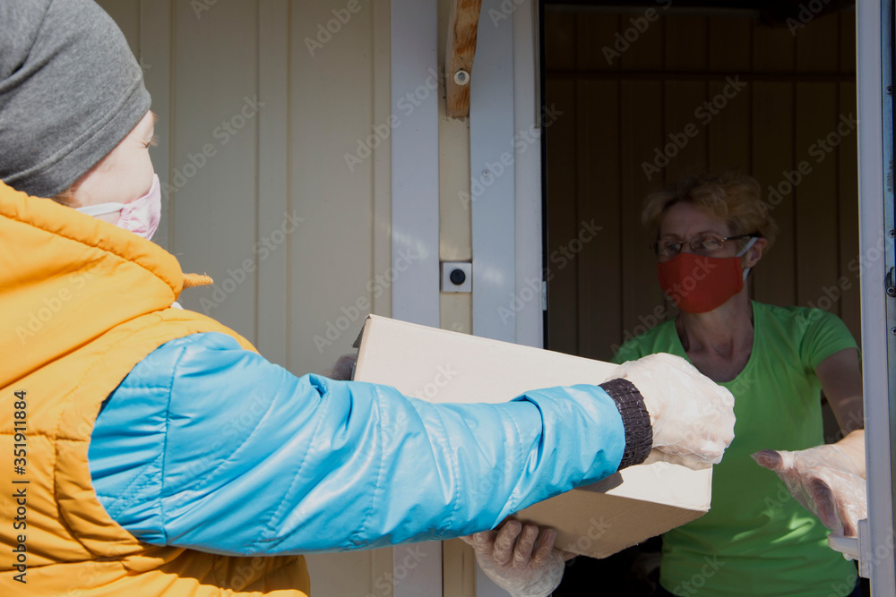 a volunteer passes a box of products to a middle-aged woman. coronavirus. Volunteer and woman in a medical mask and gloves. distance. social help. the door is open to the country house