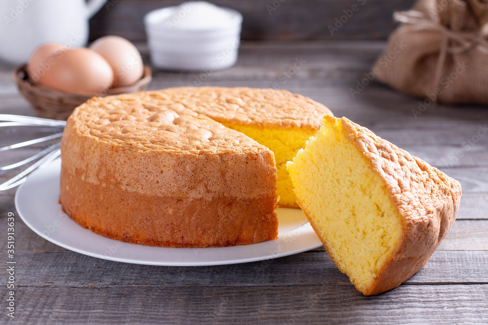 Homemade round sponge cake or chiffon cake on white plate so soft and delicious with ingredients: eggs, flour, milk on wood table Homemade round sponge cake or chiffon cake on white plate so soft and delicious with ingredients: eggs, flour, milk on wood table