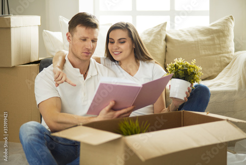 Cute couple unpacking cardboard boxes in their new home, sitting on the floor and looking at a family album