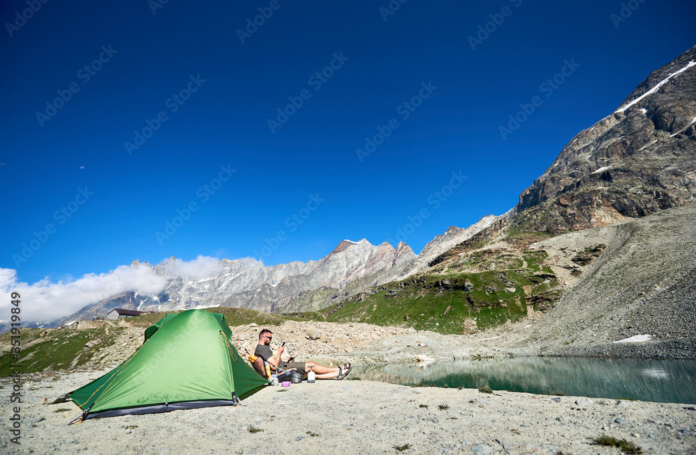 Male traveler sitting by tourist tent with majestic rocky hills on background. Young man using smartphone and enjoying rest in mountains under blue sky. Concept of travelling, hiking and camping.