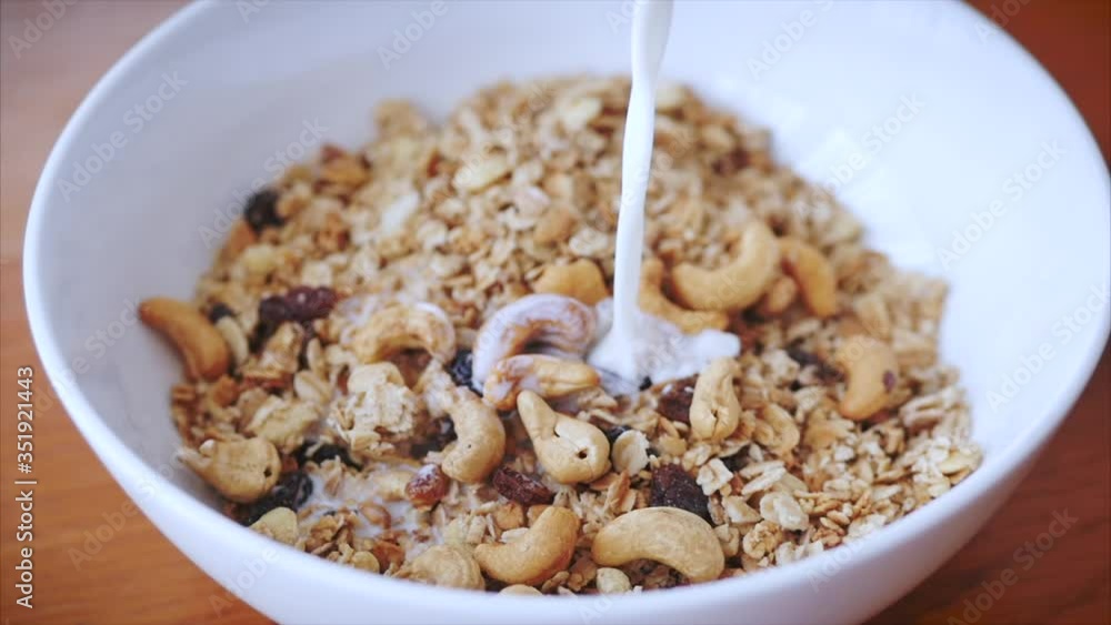 The concept of breakfast, a healthy diet, granola with cashew nuts and slices of raspberries and raisins. Slow motion milk pouring in bowl of muesli breakfast cereals. Healthy food The concept of breakfast, a healthy diet, granola with cashew nuts and slices of raspberries and raisins. Slow motion milk pouring in bowl of muesli breakfast cereals. Healthy food