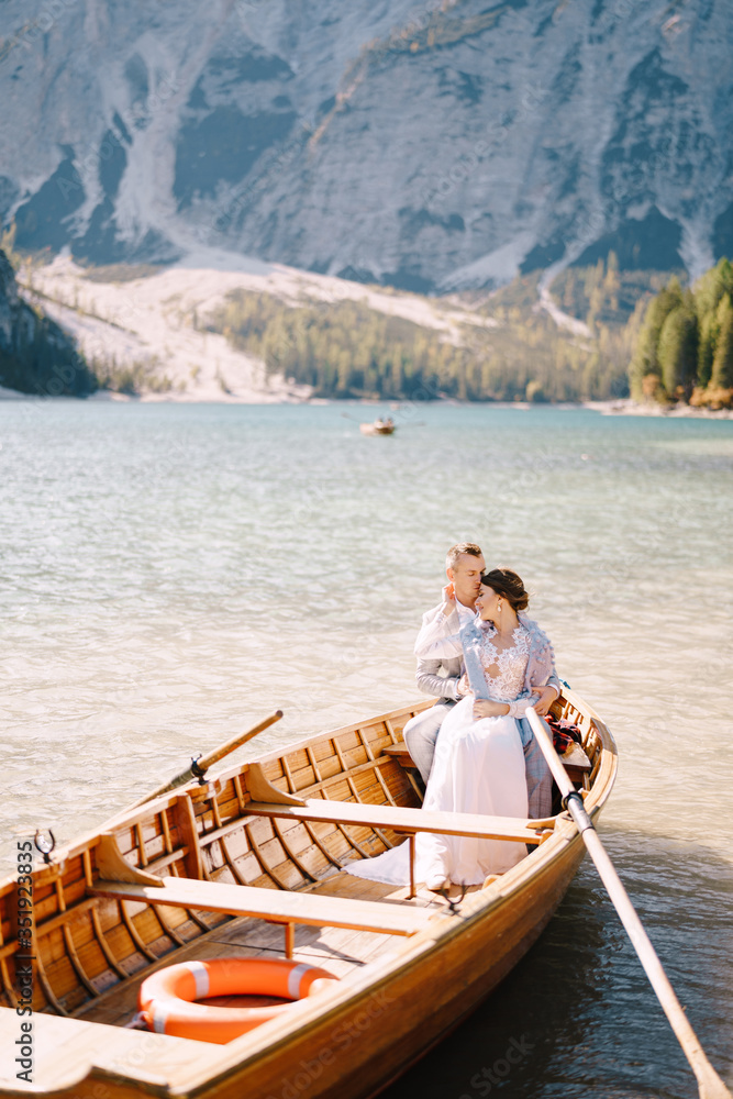 A wedding couple is sitting in a wooden boat on the Lago di Braies in Italy. Newlyweds in Europe, on Braies lake, in the Dolomites. The groom hugs the bride.