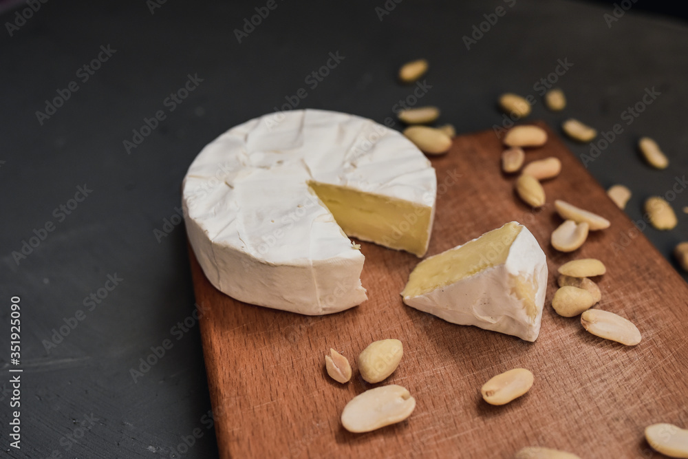 Camembert round cheese and a slice lie on a wooden board. grey matte concrete background. peanut Camembert round cheese and a slice lie on a wooden board. grey matte concrete background. peanut