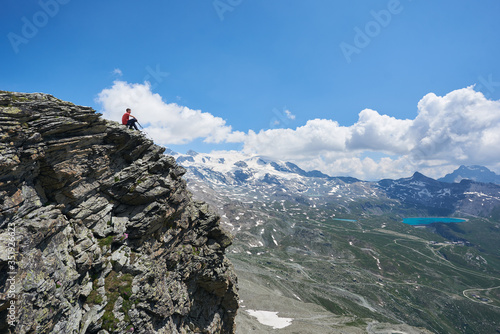 Beautiful view of traveler sitting on the edge of high rocky cliff under cloudy sky. Man tourist admiring view of mountain valley with hills and blue lake. Concept of travelling, hiking and alpinism.