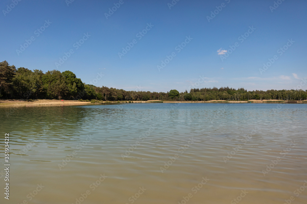 Dutch lake with a beach in the Netherlands used for recreation, swimming and sunbathing in Brabant. Calm water, greenery, sand and a blue sky. Relaxation