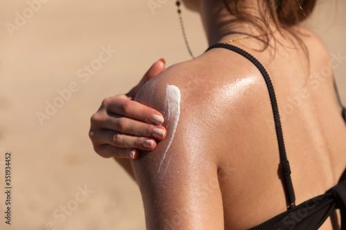 A young woman applying sun cream or sunscreen on her tanned shoulder to protect her skin from the sun. Shot on a sunny day with blurry sand in the background. photo