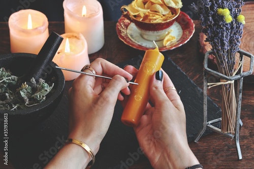 Wiccan witch carving symbols and sigils onto yellow gold color candlestick at her altar. Holding candle in hands. Witchy elements in slightly blurred background, like burning candles and dried flowers photo