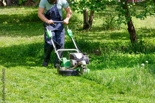 A worker mows a green lawn with a petrol mower in a spring garden.