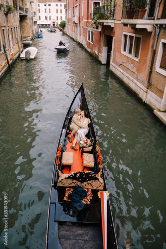 Italy wedding in Venice. A gondolier rolls a bride and groom in a classic wooden gondola along a narrow Venetian canal. Newlyweds are sitting in a boat on the background of ancient buildings.