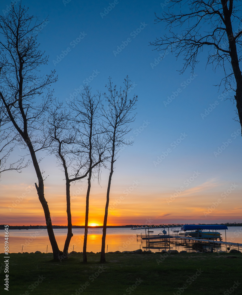 Sunset over the lake with trees, dock, lifts, and a boat in the foreground in silhouette.