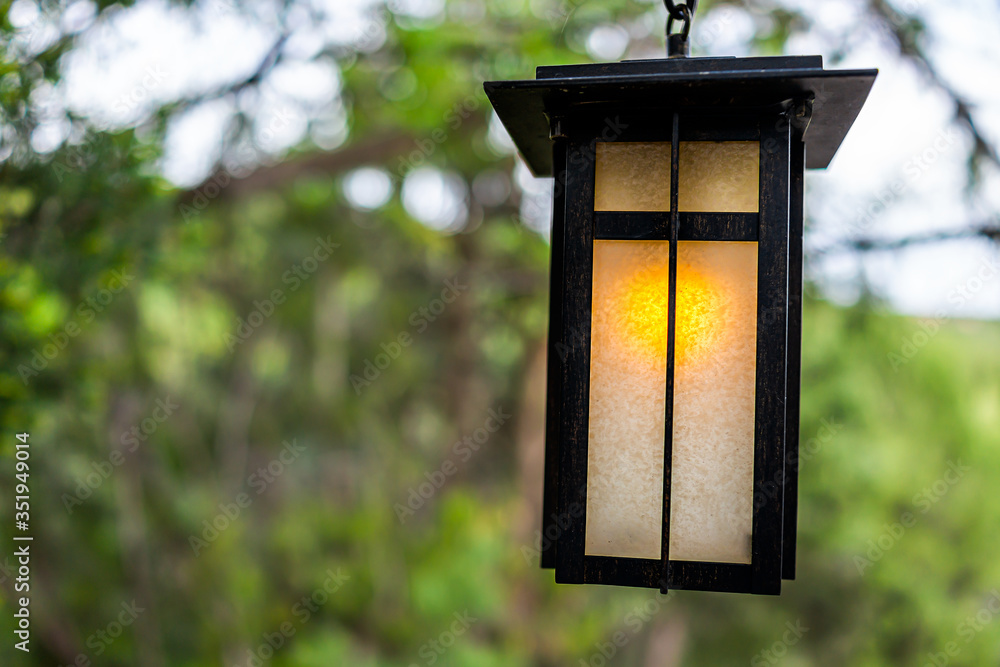 Japanese garden closeup of yellow illuminated hanging lantern in traditional style with blurry bokeh background of green trees in park at evening