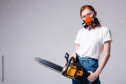 Red-haired girl in white T-shirt in construction respirator and goggles holds a chainsaw. Advertising banner for a hardware store