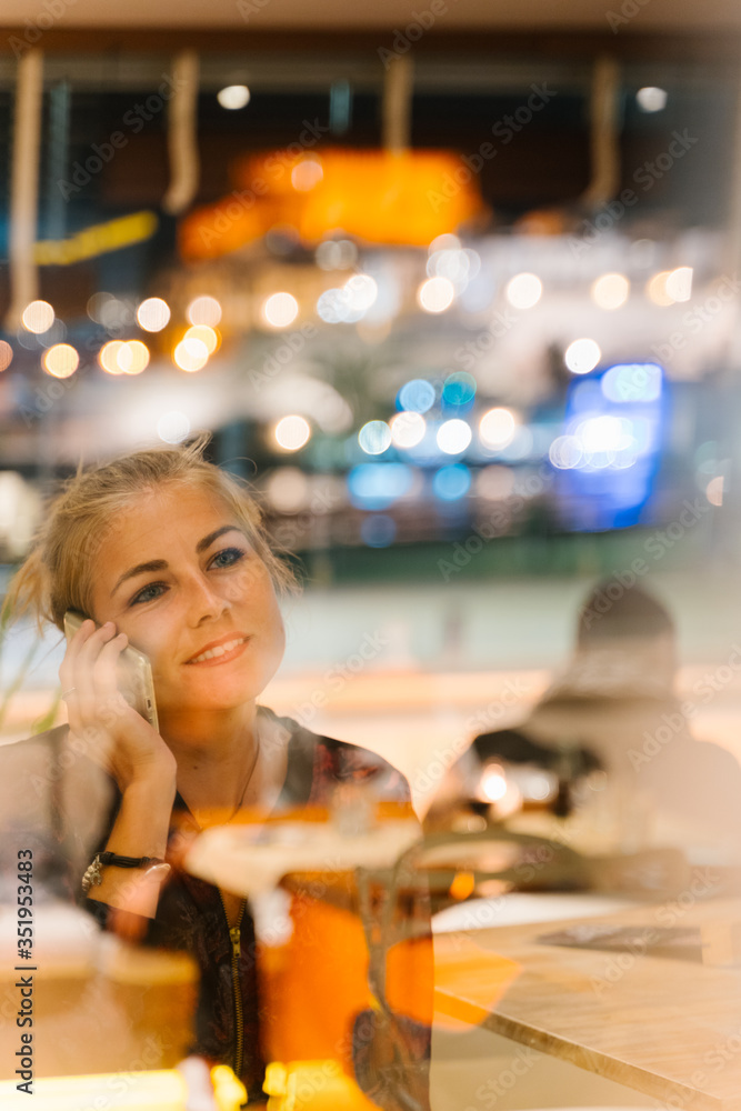 Blonde woman in a restaurant using her mobile phone and the street lights reflected in the glass at night