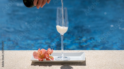 Waiter pours champagne into a glass with bubbles and foam on swimming pool or sea blurred background. Sparkling white wine on tray with flower. Cold champagne, luxury alcohol drink, romantic vacation. photo