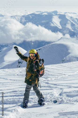Girl  bright helmet and backpack on a snowboard admires snow-capped mountain peaks and clouds which hanging low overhead. She is sending the greetings to camera with happy smile and with hands up. 