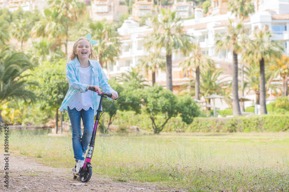 Active little toddler girl riding scooter on road in park outdoors on summer day.  Summer time fun