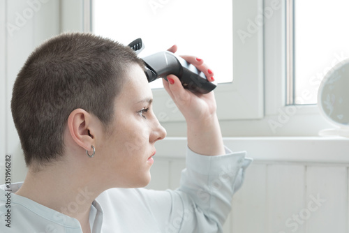 A young brunette cuts her own hair in front of a mirror. A hair clipper is in my hand. Home haircut during the quarantine. Kovid, the virus. photo