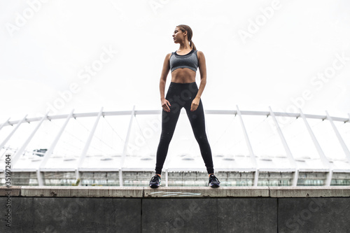 Beautiful strong and sporty young woman in sportswear stands in confident pose on the rooftop. Outdoors workout concept