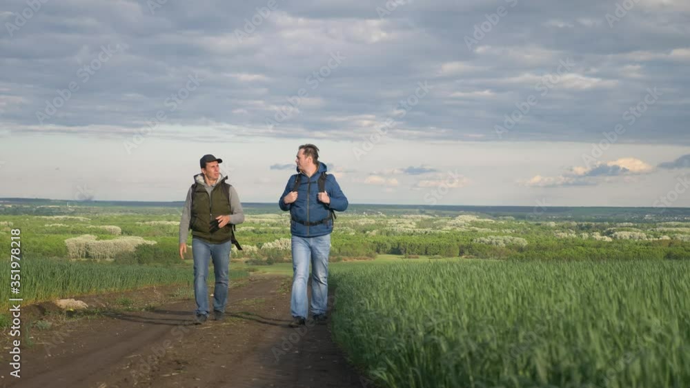 teamwork travel. two man hikers with backpacks walk lifestyle along trail next to a field with green grass in nature. concept travel adventure hiking healthy. two tourists lifestyle walk walking teamwork travel. two man hikers with backpacks walk lifestyle along trail next to a field with green grass in nature. concept travel adventure hiking healthy. two tourists lifestyle walk walking