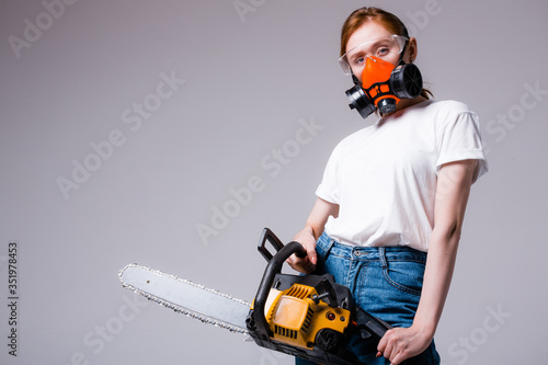 The girl in goggles and a red professional respirator holds a chainsaw in her hands and looks at the camera.