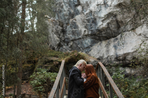 Red girl and blond guy gently hold each other's hands in the woods against the backdrop of a cliff