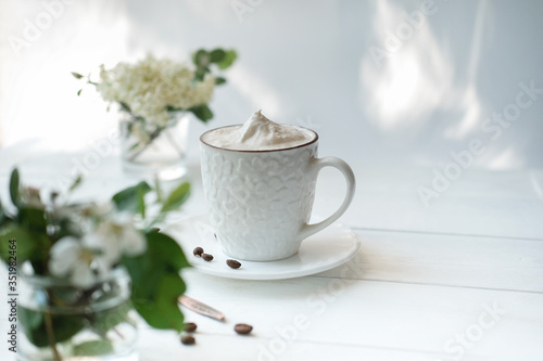 coffee cup with whipped cream on a white wooden table. cappuccino in a white cup. trendy morning light. flowers and coffee beans. good morning concept.