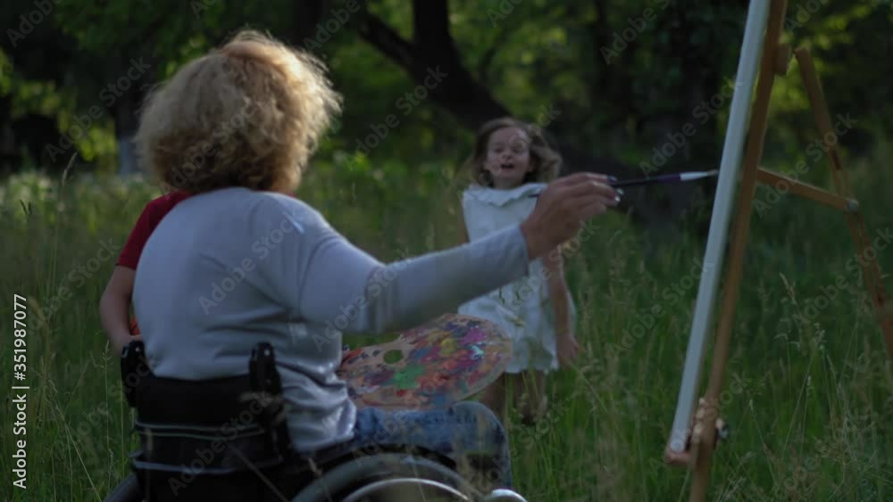 Disabled mother working on a new painting of nature using easel, spending time with her kids boy and girl playing ball around her in backyard or park Disabled mother working on a new painting of nature using easel, spending time with her kids boy and girl playing ball around her in backyard or park