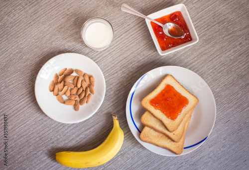 Top view of a meal made with a toast with strawberry marmalade, milk, a banana and a plate of almonds over the table