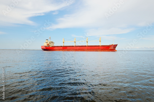 Large red bulk carrier (cargo ship) with a cranes sailing in an open sea from Europoort. Clear blue sky with cirrus clouds. Rotterdam, Netherlands. Global communications, logistics, industry theme photo