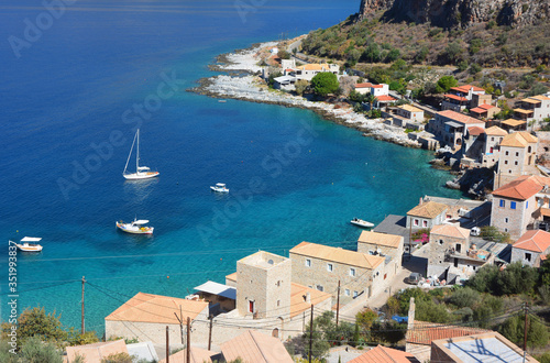 Three white yachts in the background of the beautiful turquoise Mediterranean Sea. In the foreground are several old stone houses of Peloponnese, Greece.
