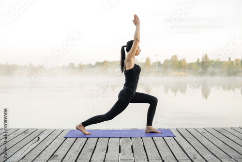 Young woman practicing yoga outdoors. Young attractive yogi brunette standing on yoga mat in warrior yoga posture. Harmony and meditation concept. Healthy lifestyle.