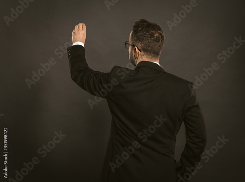 Smart businessman writes in chalk on a black wall. Rear view of white collar worker in glasses wearing black formalwear while standing on grey background. Copy space for text at left side
