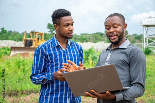 two african agricultural businessmen, discussing using a laptop on a plot of land