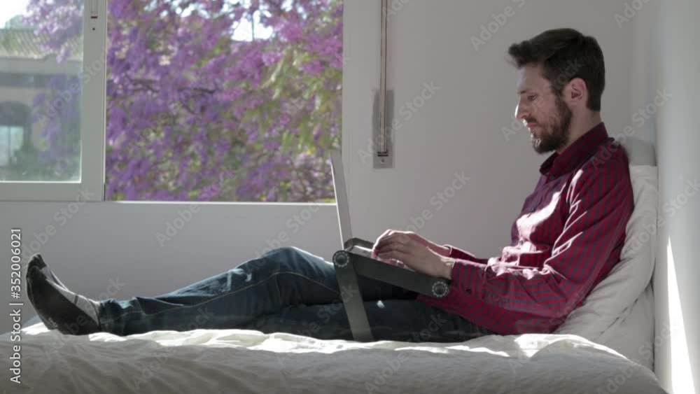 Young adult studying and working at home with his laptop using a computer stand at bed in front of the window Young adult studying and working at home with his laptop using a computer stand at bed in front of the window