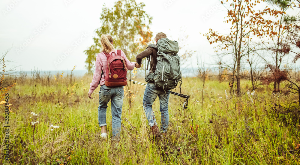 Rear view of a traveling couple of backpacker wading outdoors through an autumn field holding hands with hiking poles. Hiking concept Rear view of a traveling couple of backpacker wading outdoors through an autumn field holding hands with hiking poles. Hiking concept