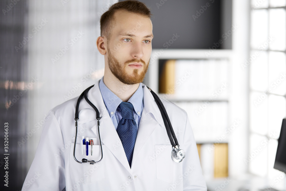 Red-bearded doctor standing straight in clinic near his working place. Portrait of physician. Medicine and healthcare concept Red-bearded doctor standing straight in clinic near his working place. Portrait of physician. Medicine and healthcare concept