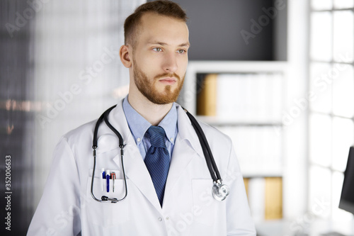 Red-bearded doctor standing straight in clinic near his working place. Portrait of physician. Medicine and healthcare concept