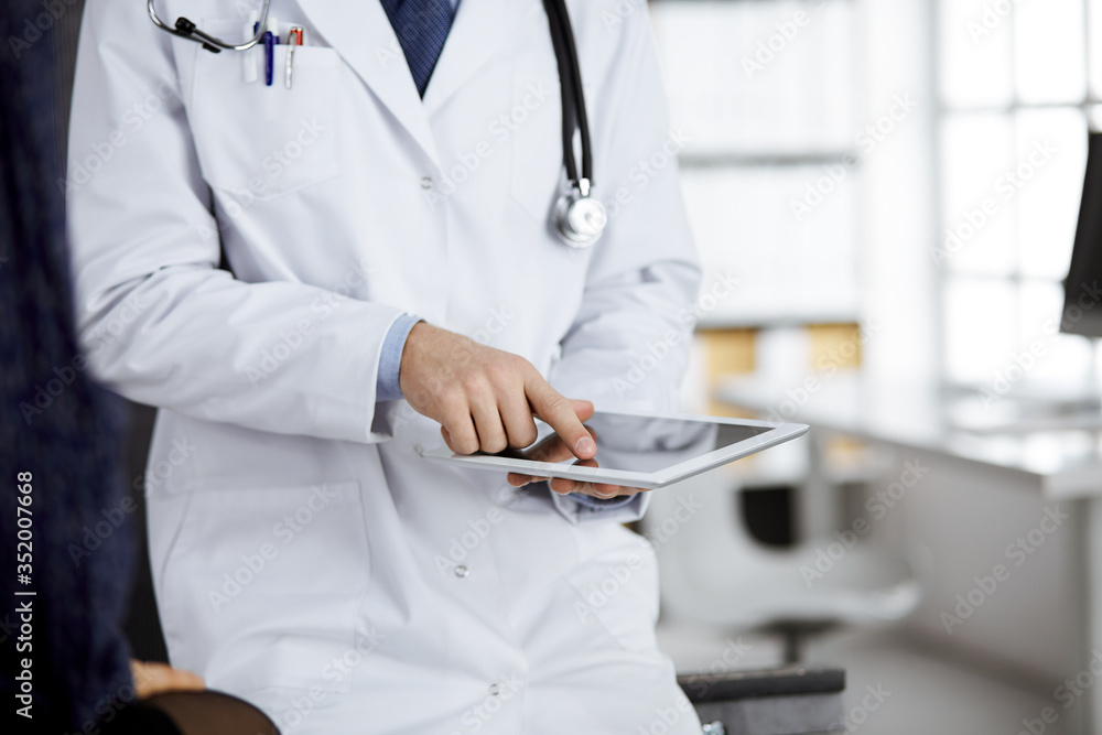 Unknown red-bearded doctor and patient woman discussing current health examination while sitting and using tablet computer in clinic, close-up. Medicine concept Unknown red-bearded doctor and patient woman discussing current health examination while sitting and using tablet computer in clinic, close-up. Medicine concept